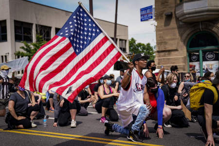 A protester wearing a Philadelphia 76ers jersey kneels while holding an American flag during a demonstration in Philadelphia, Pa. on June 3, 2020. 